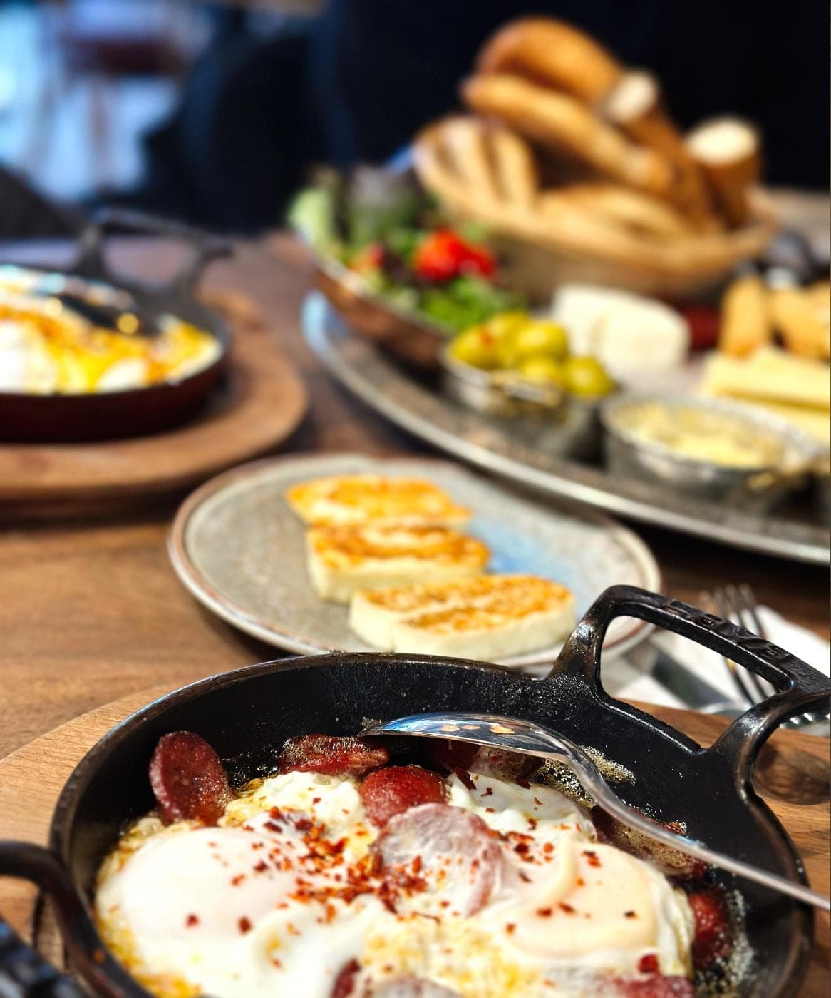 Sucuk and eggs in a cast-iron pan, with grilled halloumi and a Turkish breakfast spread behind