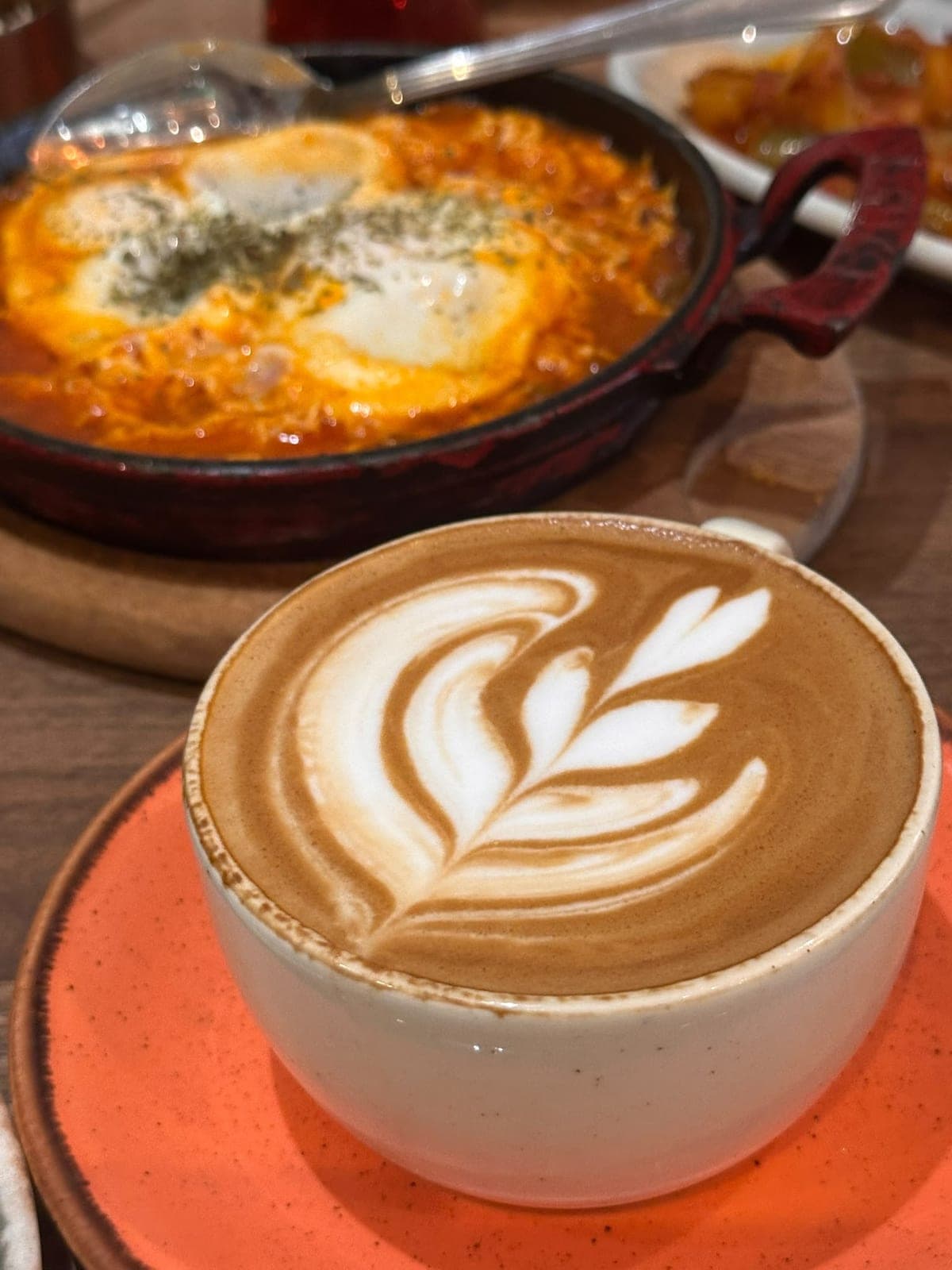 A cappuccino with a swirled leaf pattern, shakshuka bubbling behind