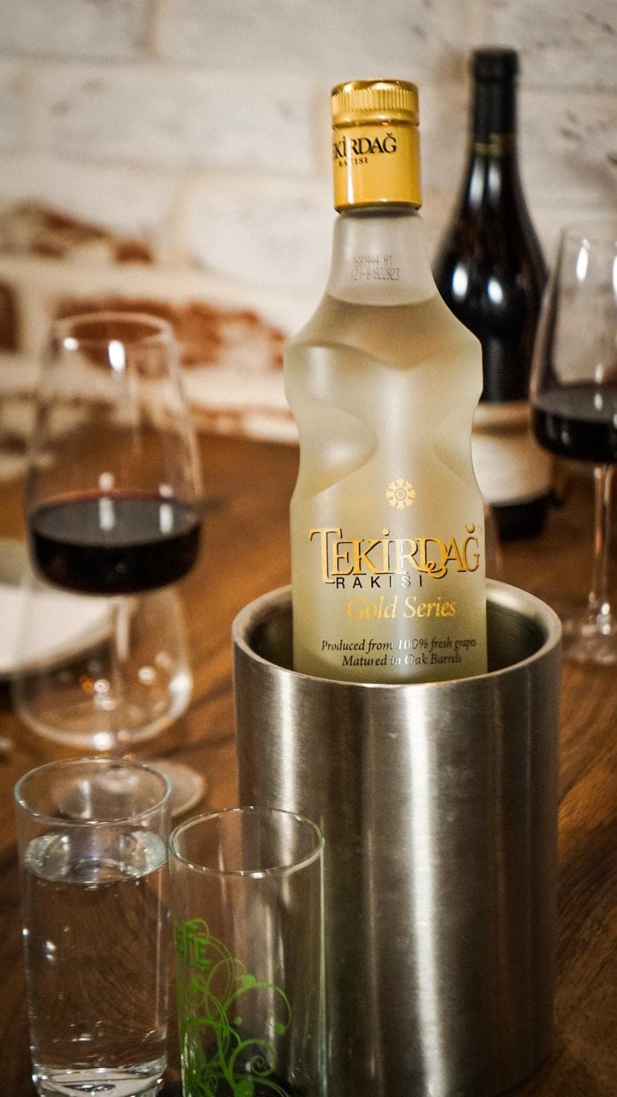 A frosted bottle of Tekirdağ rakı chilling in a steel bucket, red wine and glasses on the table